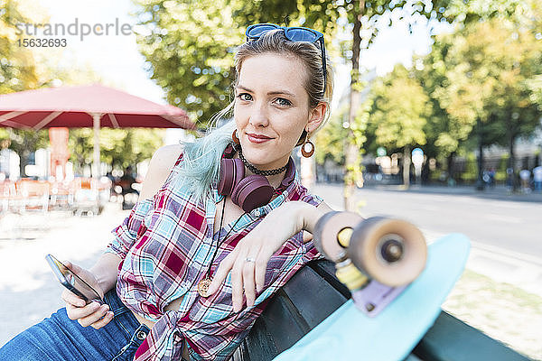 Porträt einer lächelnden jungen Frau auf einer Bank sitzend mit Skateboard und Smartphone