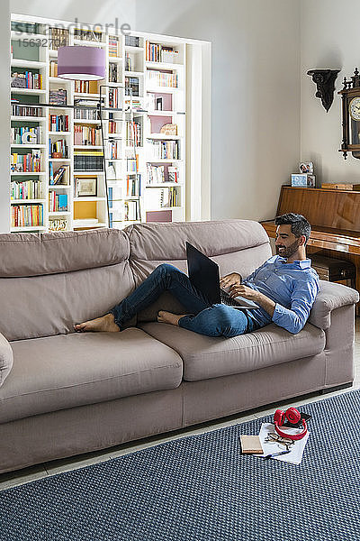 Lächelnder junger Mann  der zu Hause auf der Couch liegt und seinen Laptop benutzt