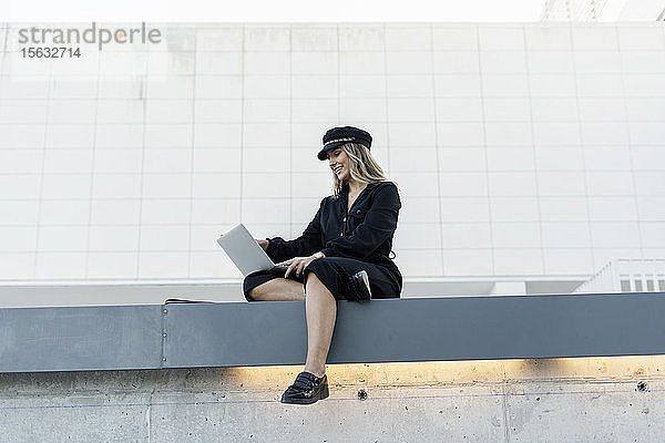 Junge blonde Geschäftsfrau mit schwarzer Matrosenmütze und Laptop  die an einer Wand sitzt