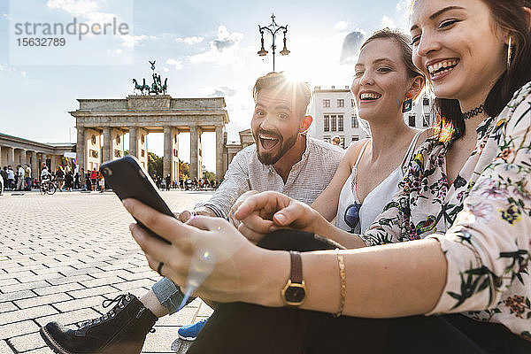 Drei lachende Freunde sitzen am Brandenburger Tor und schauen auf ein Smartphone  Berlin  Deutschland