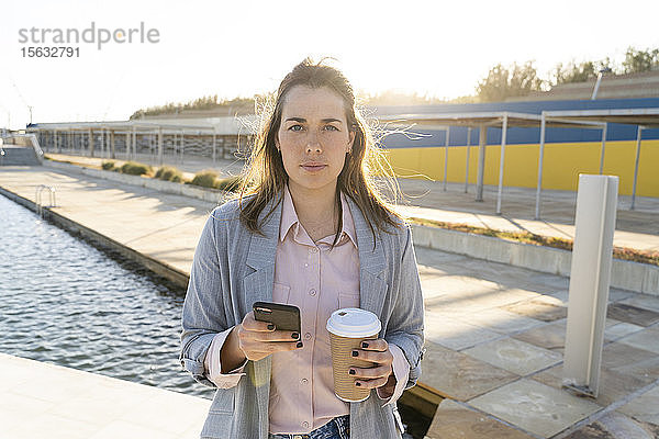 Porträt einer seriösen jungen Frau mit Handy und Kaffee zum Mitnehmen