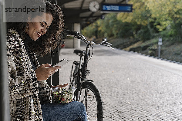 Lächelnde Frau mit Fahrrad beim Mittagessen und mit einem Smartphone auf dem Bahnsteig  Berlin  Deutschland