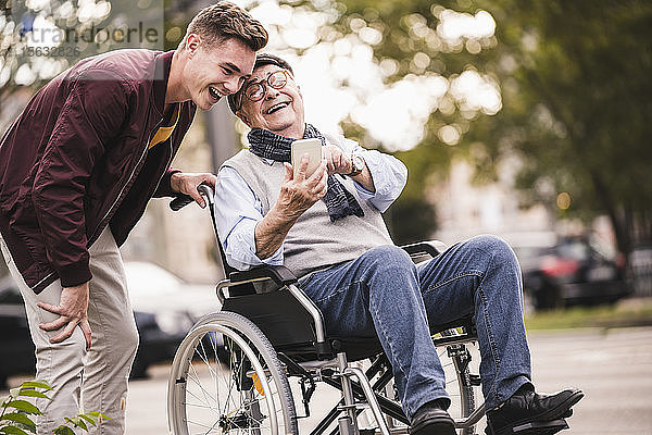 Lachender älterer Mann im Rollstuhl und sein erwachsener Enkel schauen gemeinsam auf Smartphone und haben Spaß