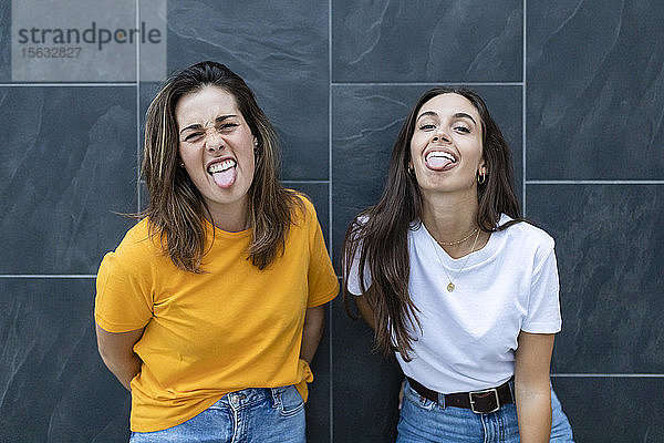Zwei Freundinnen  die an einer Wand lehnen und ihre Zungen herausstrecken
