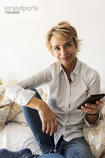 Porträt einer lächelnden reifen Frau  die zu Hause auf dem Bett sitzt und ein Smartphone hält