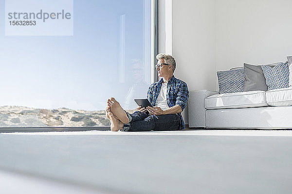 Lässiger Geschäftsmann sitzt in modernem Haus vor dem Fenster und hält Tablet