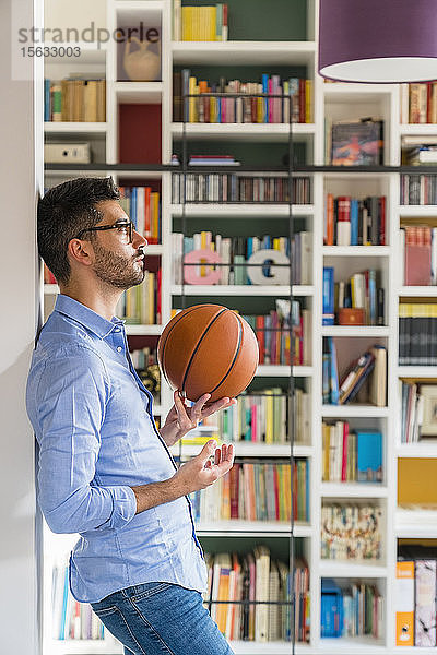 Nachdenklicher junger Mann mit Basketball  der zu Hause vor einem Bücherregal steht