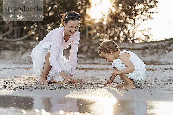 Glückliche Mutter mit Tochter spielt mit Sand am Strand