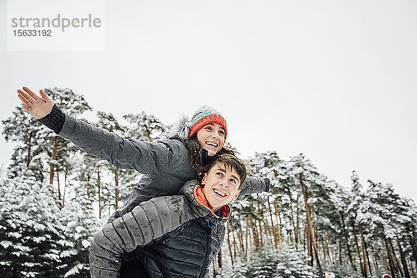 Porträt eines lächelnden jungen Mannes  der seine glückliche Freundin im Winterwald Huckepack nimmt