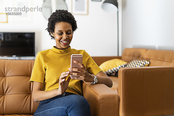 Lächelnde junge Frau sitzt zu Hause auf der Couch und benutzt ein Smartphone