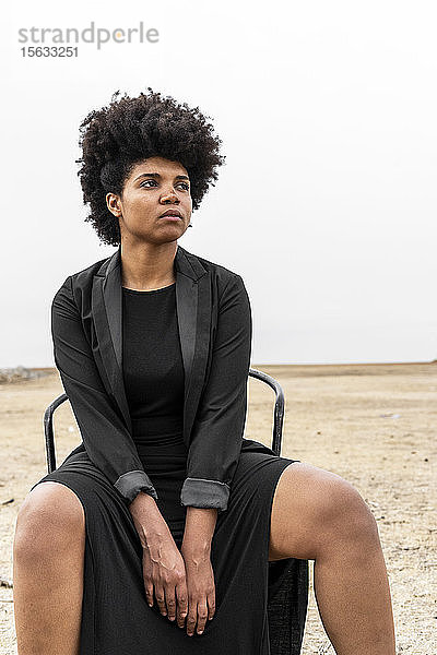 Porträt einer schwarz gekleideten jungen Frau  die in trostloser Landschaft sitzt und in die Ferne schaut