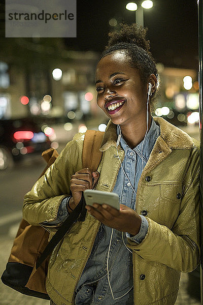 Porträt einer glücklichen jungen Frau mit Kopfhörer und Smartphone in der Stadt bei Nacht  Lissabon  Portugal