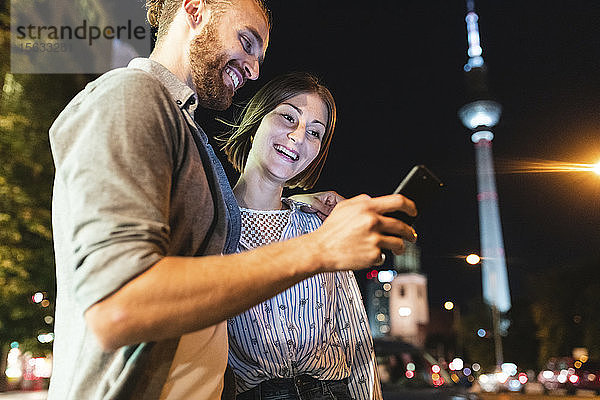 Glückliches Paar benutzt nachts in der Stadt Smartphones  Fernsehturm im Hintergrund  Berlin  Deutschland