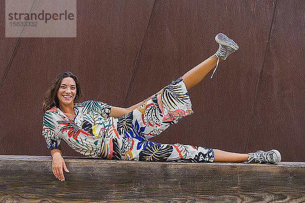 Porträt einer lächelnden jungen Frau  die auf einer Holzbank liegt und ihr Bein hebt