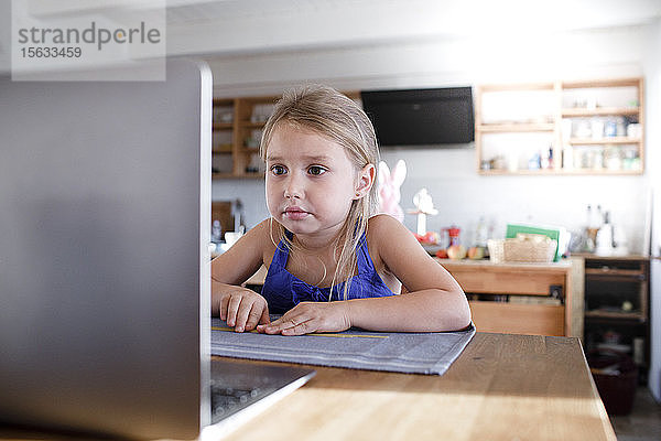 Porträt eines kleinen Mädchens  das zu Hause in der Küche sitzt und auf den Laptop starrt