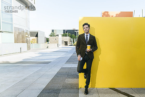 Geschäftsmann mit Kaffee zum Mitnehmen und Telefon neben der gelben Wand im städtischen Geschäftsviertel  Madrid  Spanien