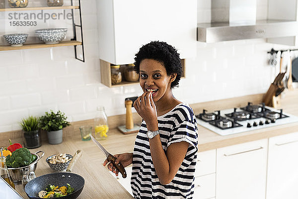 Porträt einer lächelnden jungen Frau  die zu Hause in der Küche kocht und Gemüse probiert