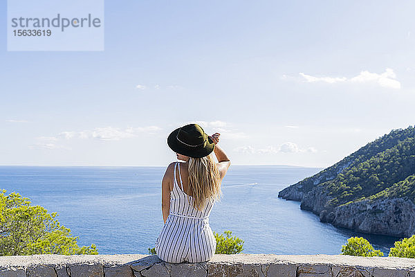 Junge Frau sitzt auf einer Mauer  Hand auf dem Hut  Aussichtspunkt auf Ibiza