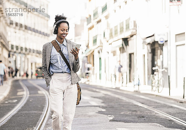 Lächelnde junge Frau mit Kopfhörer und Mobiltelefon in der Stadt unterwegs  Lissabon  Portugal