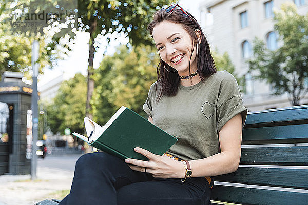 Porträt einer lächelnden jungen Frau  die mit einem Buch auf einer Bank sitzt
