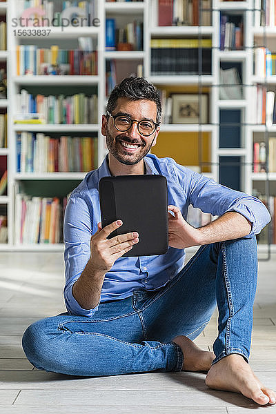Porträt eines barfüssigen jungen Mannes  der vor Bücherregalen auf dem Boden sitzt und ein digitales Tablett benutzt