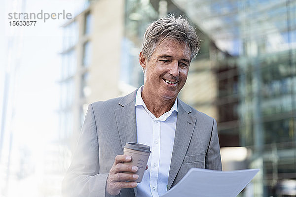 Lächelnder reifer Geschäftsmann mit Kaffee zum Mitnehmen bei der Durchsicht von Dokumenten in der Stadt