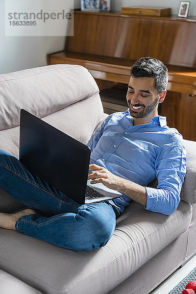 Lächelnder junger Mann  der zu Hause auf der Couch sitzt und seinen Laptop benutzt