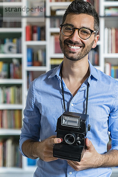 Porträt eines glücklichen jungen Mannes mit Sofortbildkamera vor einem Bücherregal stehend