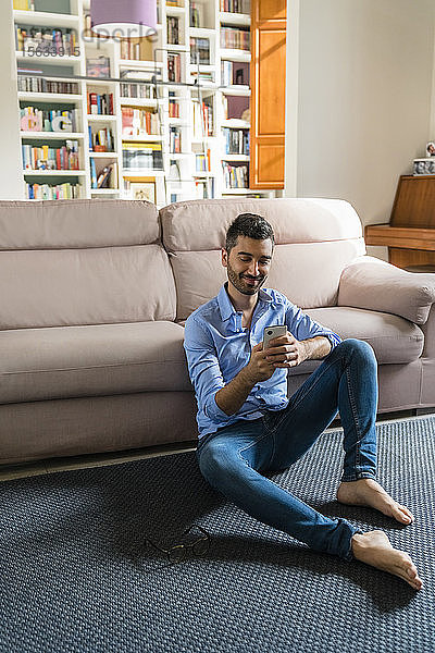 Porträt eines lächelnden jungen Mannes  der zu Hause auf dem Boden des Wohnzimmers sitzt und ein Smartphone benutzt