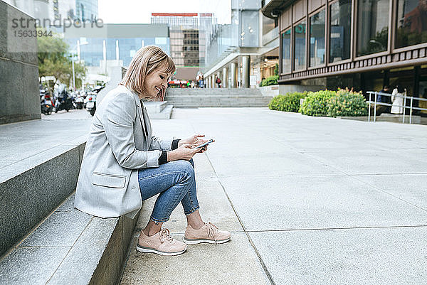 Lächelnde Frau sitzt auf einer Treppe in der Stadt und benutzt ihr Handy