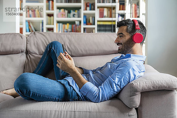 Lächelnder junger Mann  der zu Hause auf der Couch liegt und ein Smartphone und drahtlose Kopfhörer benutzt