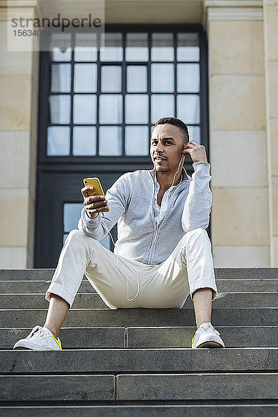 Porträt eines Geschäftsmannes  der in der Stadt auf einer Treppe sitzt und mit Kopfhörern und Smartphone Musik hört