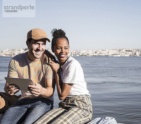 Glückliches junges Paar sitzt mit einer Tablette am Wasser  Lissabon  Portugal