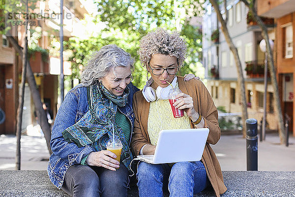 Ältere Mutter und ihre erwachsene Tochter nutzen Laptop und trinken Säfte in der Stadt