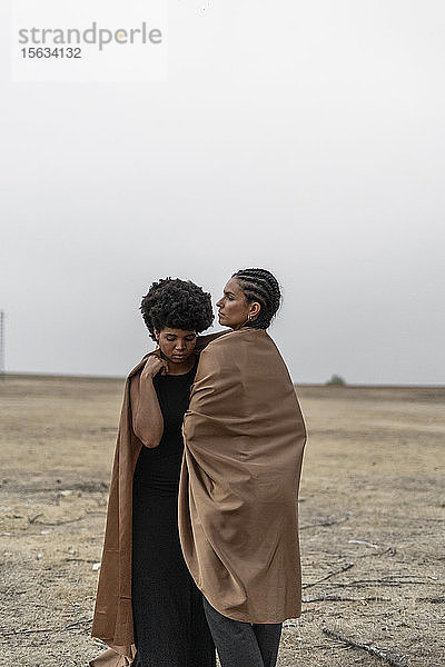 Zwei Frauen stehen in trostloser Landschaft und teilen sich eine Decke