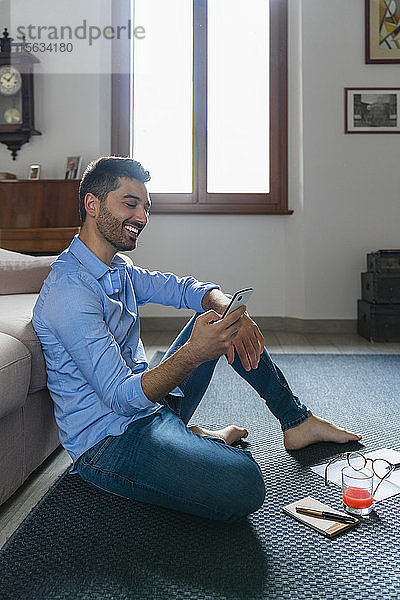 Porträt eines lächelnden jungen Mannes  der zu Hause barfuss auf dem Boden sitzt und auf ein Smartphone schaut