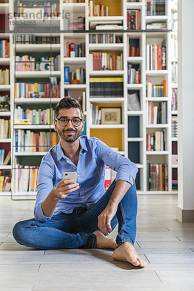 Porträt eines lächelnden jungen Mannes  der zu Hause mit seinem Smartphone vor Bücherregalen auf dem Boden sitzt