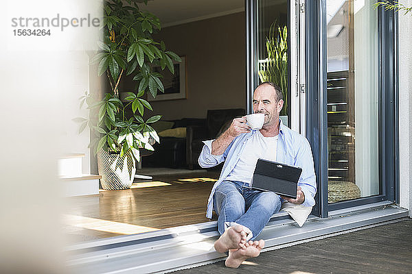 Reifer Mann sitzt zu Hause an der Terrassentür mit Tablette und Kaffeetasse