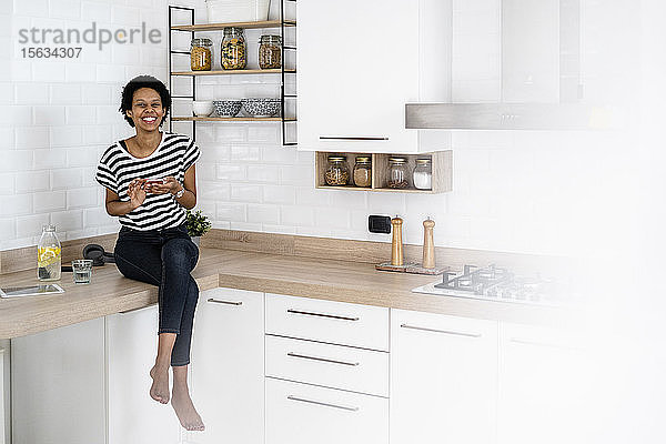 Porträt einer glücklichen jungen Frau mit Smartphone in der heimischen Küche