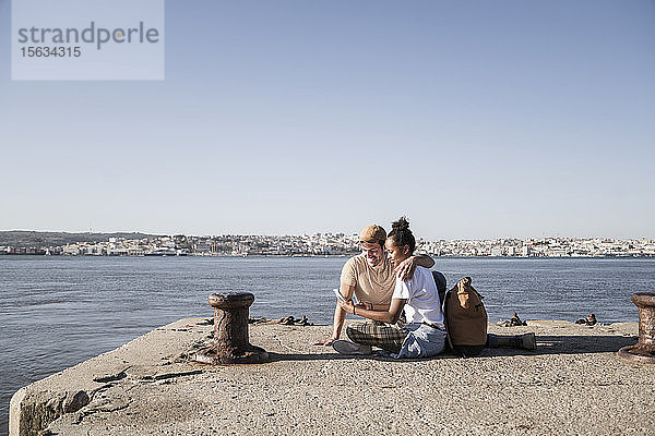 Junges Paar sitzt am Pier am Wasser und telefoniert per Handy  Lissabon  Portugal