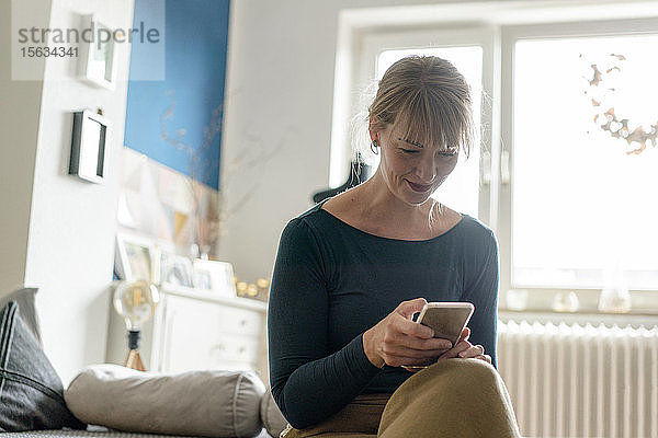 Lächelnde Frau sitzt im Wohnzimmer und benutzt ein Smartphone