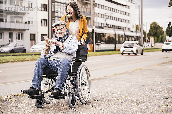 Lächelnde junge Frau schiebt glücklichen älteren Mann mit Smartphone im Rollstuhl