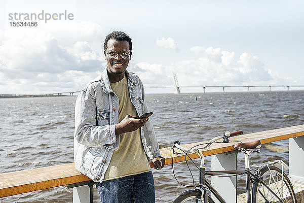 Junger Mann lehnt an der Reling am Meer und benutzt ein Smartphone