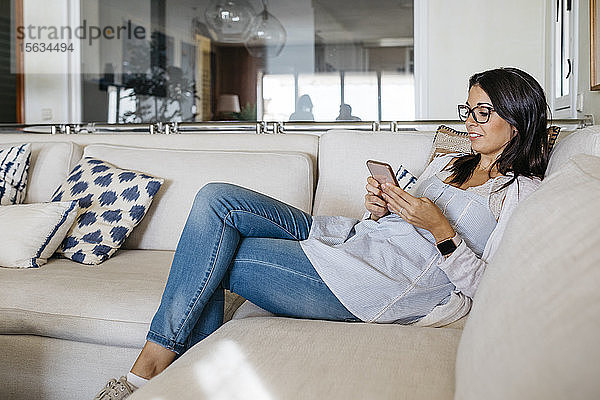 Lächelnde Frau zu Hause  die auf der Couch sitzt und auf ihr Handy schaut