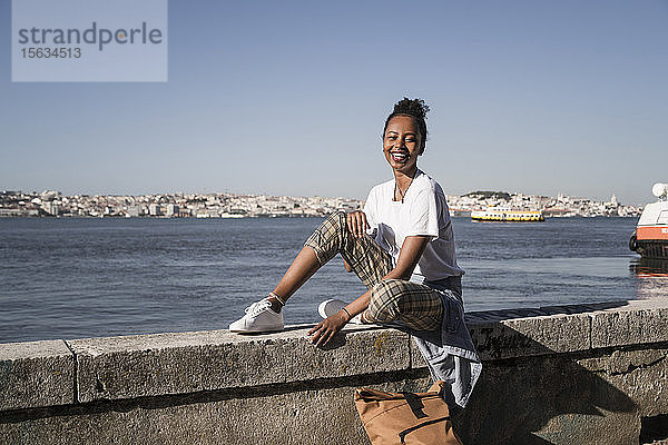 Porträt einer glücklichen jungen Frau  die auf einer Mauer am Wasser sitzt  Lissabon  Portugal