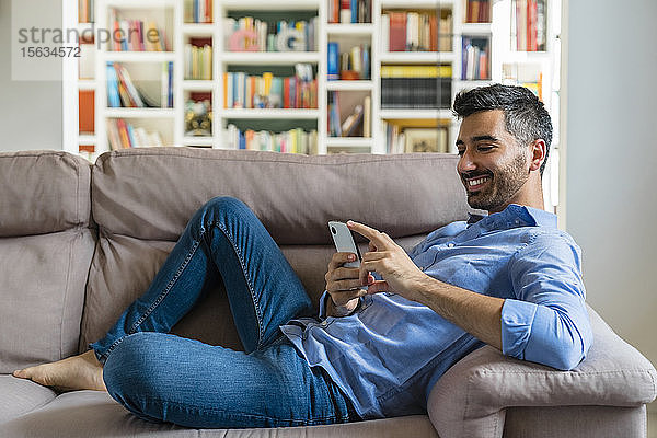 Lächelnder junger Mann liegt zu Hause auf der Couch und benutzt ein Smartphone