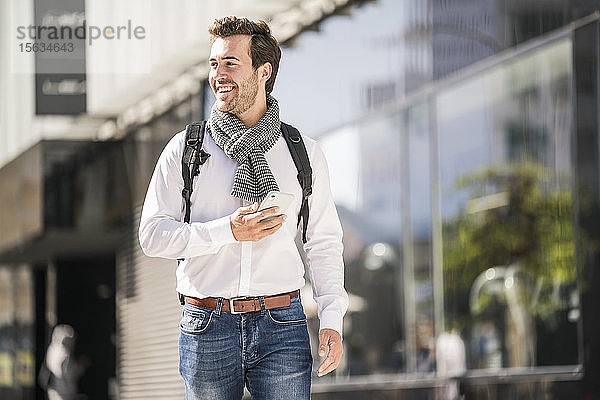 Lächelnder junger Mann mit Rucksack und Handy in der Stadt unterwegs