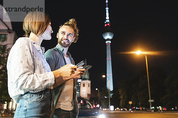 Glückliches Paar benutzt nachts in der Stadt Smartphones  Fernsehturm im Hintergrund  Berlin  Deutschland