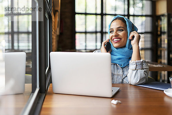 Geschäftsfrau  die in einem Café einen türkisfarbenen Hijab trägt und mit ihrem Laptop arbeitet  am Telefon