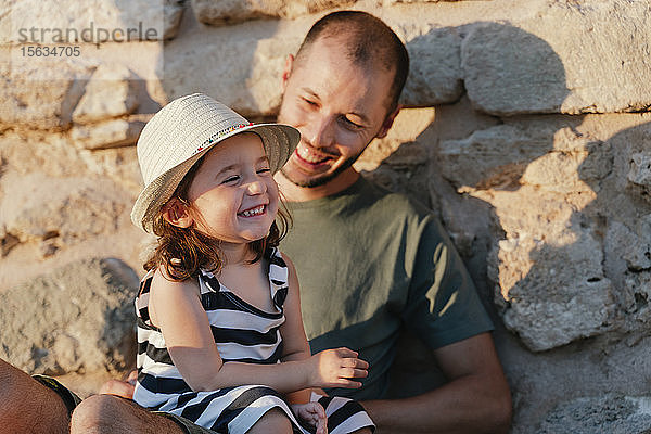 Porträt eines glücklichen kleinen Mädchens  das bei Sonnenlicht auf dem Schoss seines Vaters sitzt
